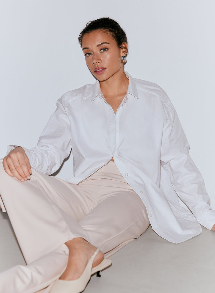 A woman with dark hair wearing a white oversized shirt and sand-colored Luxe Pants, sitting on the floor. [36-170cm]