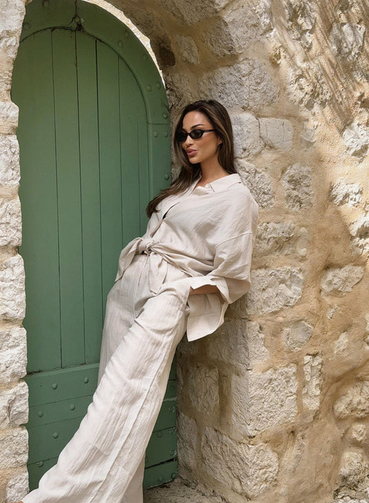 Relaxed sand Riviera Pants from aim'n worn with a matching shirt by a woman leaning against a stone wall.