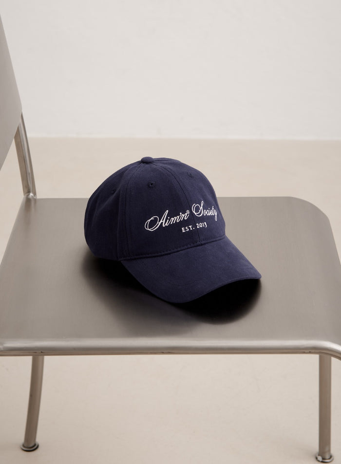 Navy blue cotton Society Cap with white aim'n embroidery resting on a grey metal chair.