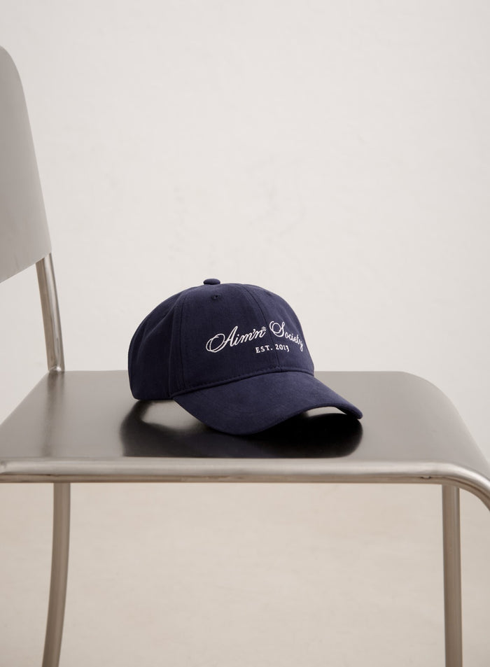Navy cotton aim'n Society Cap with white embroidered logo resting on a minimal metal chair.