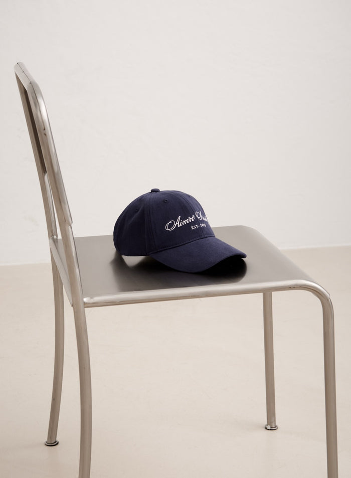 Navy cotton Society Cap featuring white embroidered aim'n logos, resting on a silver metal chair.