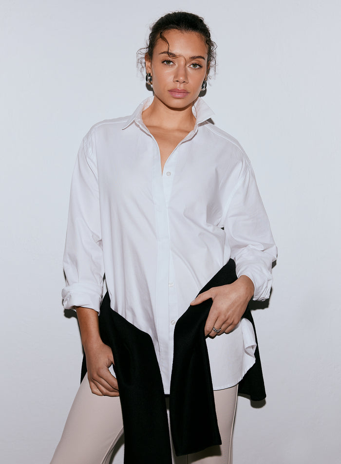 A woman wearing a white long-sleeve oxford shirt, light pants, and a black garment over her arm, looking at the camera. [M-170cm]
