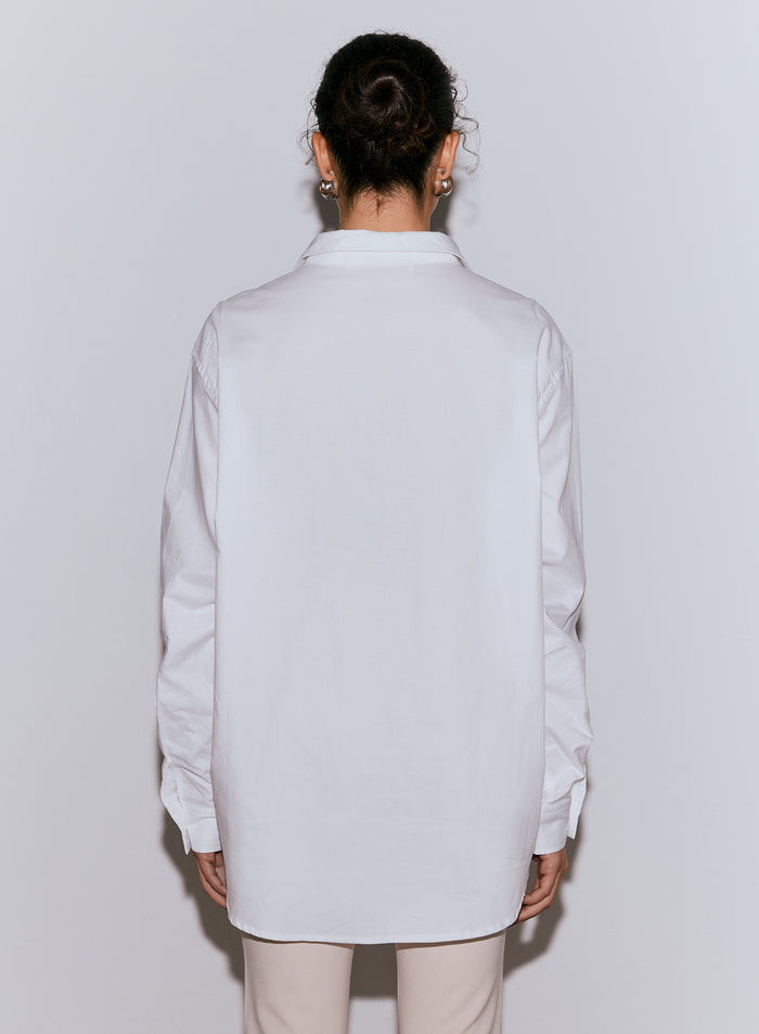 A person viewed from behind wearing a white Oxford shirt and light-colored pants, with hair in a bun and hoop earrings. [M-170cm]