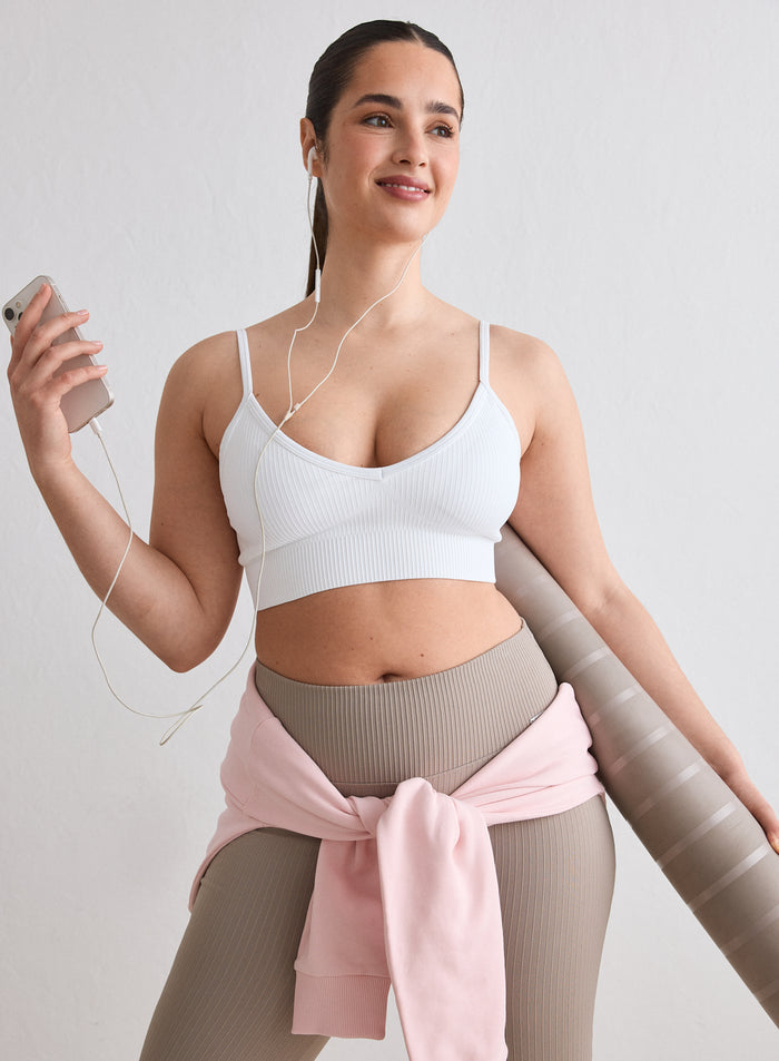 A woman wearing a White Ribbed Seamless Deep Cut Bra from aim'n, holding a yoga mat and a smartphone [L-174cm]