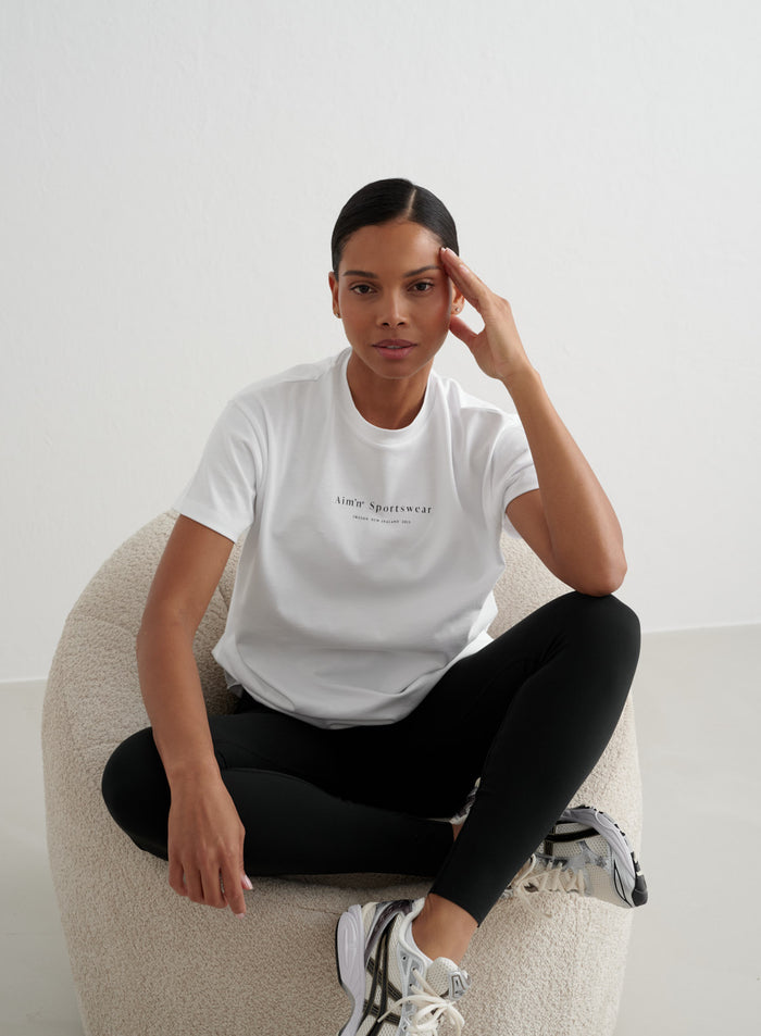 White Serif Boyfriend T-shirt from aim'n with black logo, worn by a woman sitting in a plush round chair.