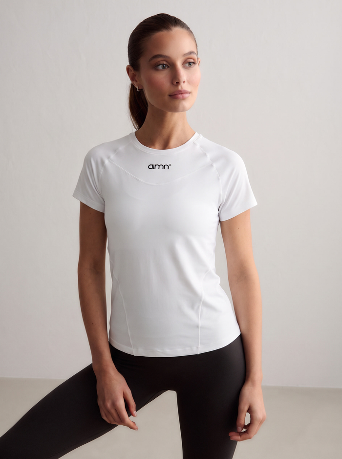 A model wearing the white Soft Basic Short Sleeve with a printed aim'n logo, paired with black tights [S - 174 cm]