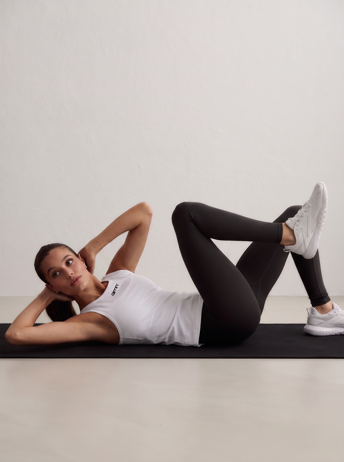 A model lying on a mat performing core exercises, wearing a white Soft Basic Singlet, black leggings and white sneakers [S - 174 cm]